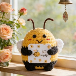 Op de afbeelding: Een gehaakte bij met een geel en zwart lichaam, die een kaart vasthoudt met de tekst "BEE happy". De bij heeft grote zwarte ogen, rode wangen en witte vleugels. Op de achtergrond perzikrozen en een raam.