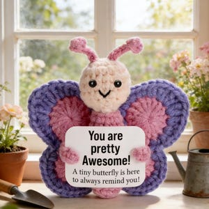 May include: A crocheted butterfly with pink, purple, and cream colors holds a sign that reads "You are pretty Awesome! A tiny butterfly is here to always remind you!" The butterfly is displayed near a window with potted plants.