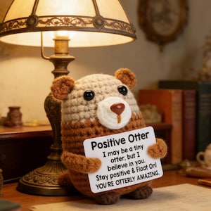 May include: A crocheted otter plushie in shades of brown and beige, holding a sign that reads "Positive Otter." The sign includes an inspirational message. The plushie has black eyes and a white muzzle.
