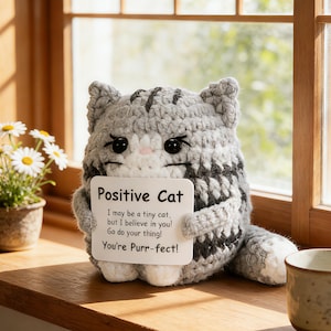 Może przedstawiać: Szydełkowa szara i biała figurka kota trzymająca znak z napisem "Positive Cat". Znak zawiera frazy "I may be a tiny cat, but I believe in you! Go do your thing! You're Purr-fect!". Kot siedzi na drewnianej powierzchni.