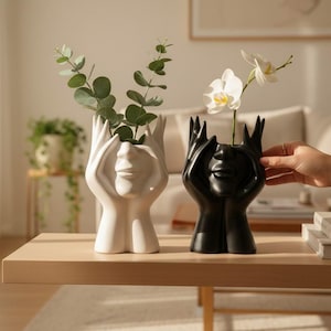Può includere: Due vasi in ceramica a forma di volto con mani, uno bianco e uno nero. Il vaso bianco contiene eucalipto verde, e il vaso nero contiene orchidee bianche. I vasi sono su un tavolo di legno chiaro.