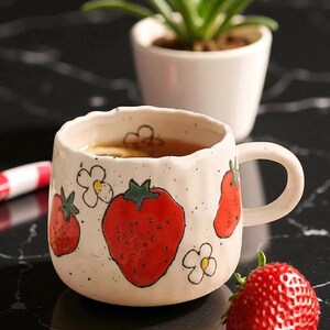 Puede incluir: Taza de cerámica blanca decorada con fresas rojas y motivos florales blancos. La taza contiene té. Una fresa fresca está colocada junto a la taza. Una pequeña planta en maceta es visible en el fondo. Capacidad aproximada de 300 ml.