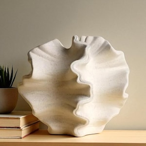 Könnte beinhalten: Eine große, cremefarbene dekorative Keramikvase mit einem wellenförmigen, muschelartigen Design. Die Vase hat eine strukturierte Oberfläche mit dezenten gesprenkelten Details. Die Skulptur steht auf einer Holzoberfläche, mit einer kleinen Topfpflanze und Büchern im Hintergrund.