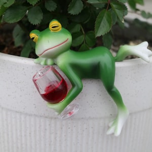 Puede incluir: Una caprichosa figura de rana verde con ojos amarillos y patas blancas, sosteniendo una copa pequeña de vino tinto. La rana está posada en el borde de una maceta blanca llena de plantas verdes. Un artículo decorativo divertido para el hogar o el jardín.