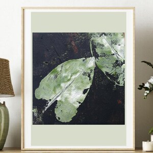 Op de afbeelding: Abstracte kunstprint met twee grote groene bladeren tegen een donkere achtergrond. De bladeren hebben een textuur, met zichtbare penseelstreken. Het kunstwerk is ingelijst in een licht houten frame, geschikt voor interieurdecoratie.