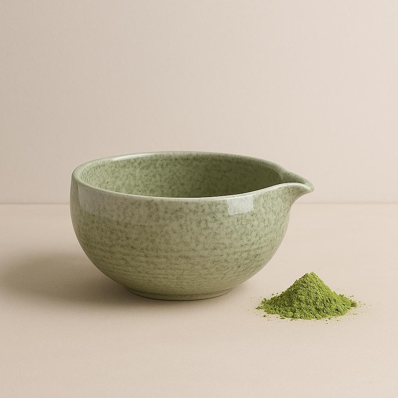 May include: A speckled green ceramic bowl with a pouring spout, next to a small pile of bright green matcha powder. The bowl has a textured surface and a rounded shape, suitable for whisking matcha.