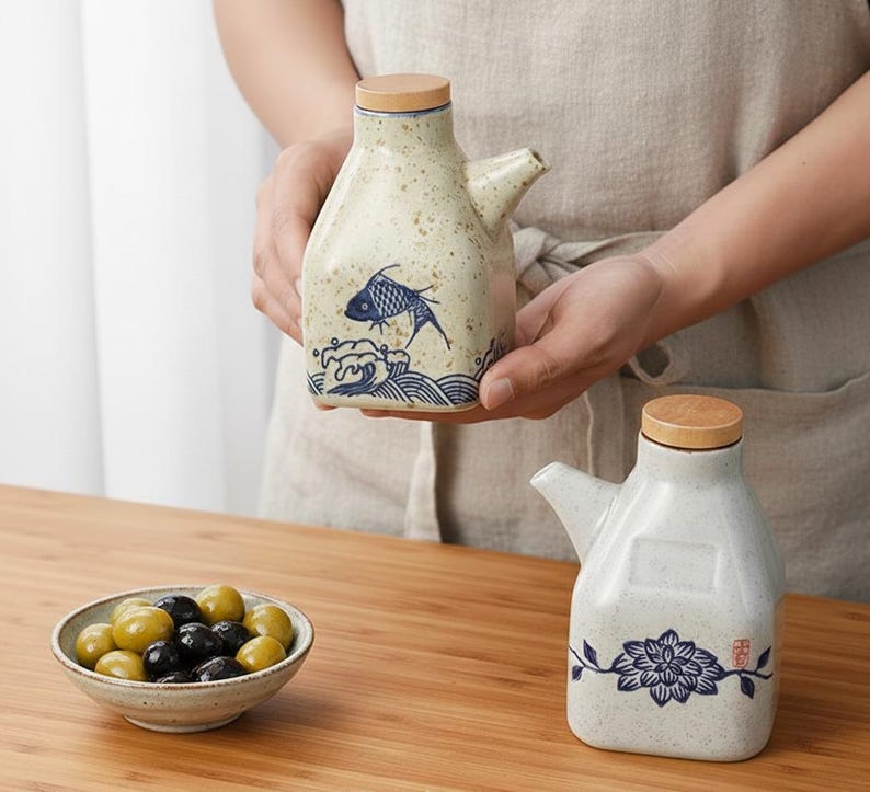 May include: Ceramic oil and vinegar dispensers with wooden lids. One dispenser features a blue fish design, the other a floral pattern. A small bowl of olives sits on a wooden surface.