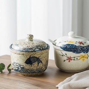 以下が含まれることがあります： 日本のデザインの蓋付きセラミックジャー2個。1つは魚と波の模様、もう1つは花と雲のモチーフです。どちらも木製の表面にあります。