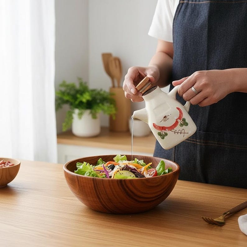 May include: A wooden salad bowl filled with fresh greens, tomatoes, and other vegetables. A ceramic oil dispenser with a chili pepper design is pouring oil onto the salad. The dispenser has a wooden lid and a handle. A wooden fork rests on the table.