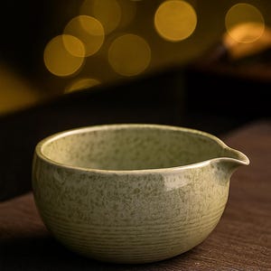 May include: A light green ceramic bowl with a spout, ideal for serving tea or other beverages. The bowl has a textured surface and a rounded shape, with a small spout for easy pouring. The bowl is sitting on a dark wooden surface.