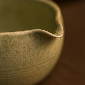 May include: Close-up of a light green ceramic bowl with a spout. The bowl has a textured surface and a curved rim. The background is a blurred brown.