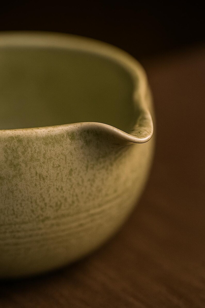 May include: Close-up of a light green ceramic bowl with a spout. The bowl has a textured surface and a curved rim. The background is a blurred brown.
