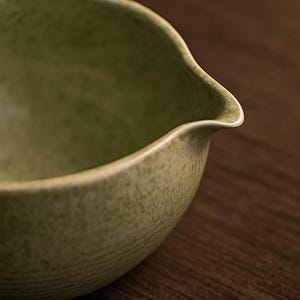May include: Close-up of a green ceramic bowl with a spout. The bowl has a textured surface and a light-colored rim. The bowl is set against a dark brown wooden background.
