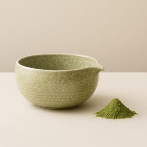 May include: A light green ceramic matcha bowl with a spout, next to a small pile of green matcha powder. The bowl has a speckled texture and a rounded shape, perfect for whisking matcha tea.