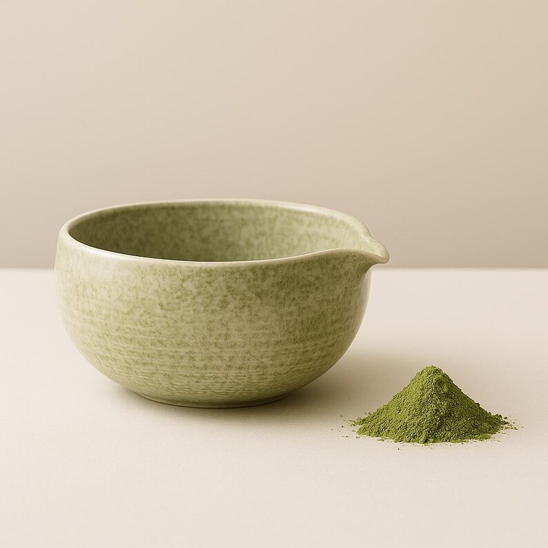 May include: A light green ceramic matcha bowl with a spout, next to a small pile of green matcha powder. The bowl has a speckled texture and a rounded shape, perfect for whisking matcha tea.