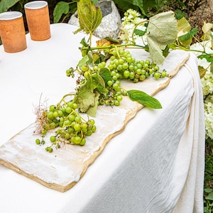 May include: A rustic table setting featuring a rectangular, off-white serving board adorned with green grapes and leaves. Four terracotta-colored cups are arranged on the white tablecloth. White flowers and greenery complete the natural aesthetic.