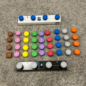 Puede incluir: Una colección de coloridas piezas de juego y dos paneles de control electrónicos. Las piezas del juego son de varios colores, incluyendo marrón, rosa, amarillo, verde, morado, rojo, azul, gris y naranja. Los paneles de control son blancos y negros con perillas azules y grises.