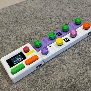 Könnte beinhalten: Ein weißes und lila elektronisches Musikgerät mit bunten Knöpfen. Das Gerät hat einen kleinen Bildschirm, der Text anzeigt, und eine Reihe runder Knöpfe in Grün, Gelb, Rosa und Orange. Das Gerät ist mit "GERANG" beschriftet.