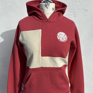 May include: Red hoodie with a cream panel on the front and a kangaroo pocket. The hoodie features a white circular logo on the right chest and a drawstring hood. The sweatshirt is displayed on a mannequin.