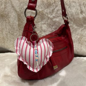 May include: Red leather handbag with a matching red strap. A heart-shaped fabric charm with pink and white stripes hangs from the bag. The charm has the word "LOVE" spelled out in colourful beads.