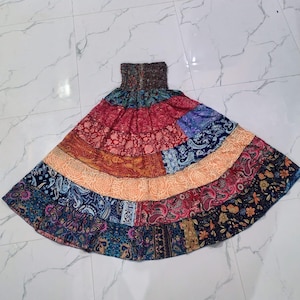 May include: A tiered maxi skirt featuring a smocked elastic waistband and a vibrant patchwork design. The skirt is composed of multiple horizontal panels displaying various floral and paisley patterns in shades of red, orange, blue, and black.