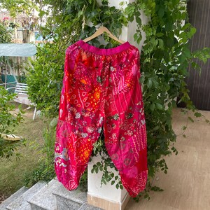 May include: A pair of loose-fitting trousers featuring a vibrant patchwork design in shades of bright pink, red, and orange. The trousers have an elasticated waistband and a multi-patterned fabric construction with floral and geometric motifs throughout.