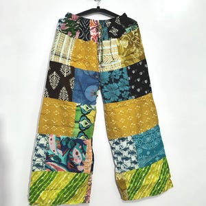 Pode incluir: Calças de perna larga com um design de patchwork composto por vários quadrados de tecido em amarelo mostarda, azul-petróleo, preto e verde-limão. A peça inclui uma cintura elástica com cordão e apresenta diversos padrões florais, geométricos e abstratos nos diferentes painéis.