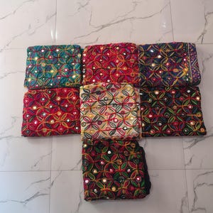 Könnte beinhalten: Sieben gefaltete Stoffquadrate mit einem sich wiederholenden kreisförmigen Blumenmuster, das mit buntem Garn und kleinen runden Spiegeln bestickt ist. Die Stofffarben umfassen Petrol, Rot, Dunkelblau, Creme und Schwarz, angeordnet in einer T-Form.