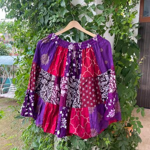 Puede incluir: Una falda hasta la rodilla con un diseño de patchwork que presenta paneles alternos de tela morada, magenta y roja oscura. La falda muestra varios patrones florales y geométricos blancos impresos en los paneles, una cintura elástica y una silueta acampanada.