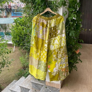May include: Wide-leg pants featuring a patchwork design in various shades of olive, lime, and chartreuse green. The garment displays a mix of floral, geometric, and botanical patterns across the fabric panels, with an elasticized waistband and drawstring.