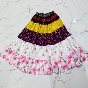 Puede incluir: Una falda larga de capas con una cintura estampada en blanco y negro, una franja amarilla con motivos de hojas blancas, una franja morada oscura con patrones florales blancos, una franja blanca con dientes de león y flores rosas.