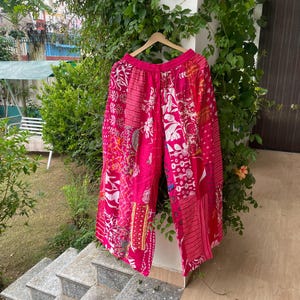 Puede incluir: Pantalones de pierna ancha con un diseño de patchwork vibrante en tonos rosa intenso, rojo y blanco. La prenda muestra varios patrones florales, geométricos y de rayas en sus paneles de tela, terminados con una cintura elástica.