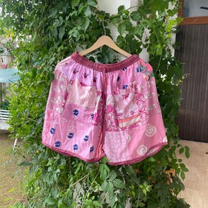 May include: A pair of pink patchwork shorts featuring a variety of floral and geometric patterns in different shades of pink, white, and blue. The shorts have an elasticized waistband with a drawstring and two front pockets, all hanging on a hanger.