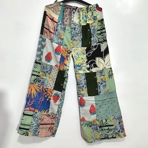 Könnte beinhalten: Weit geschnittene Hose mit einem Patchwork-Design aus verschiedenen Stoffquadraten. Die Muster umfassen rote Erdbeeren, gelbe Blumen, grünes Blattwerk und schwarze Blumenmotive auf hellblauem, weißem und olivgrünem Hintergrund.