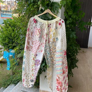 May include: Loose-fitting pants featuring a patchwork design of various floral and geometric patterns in shades of pink, red, green, and blue on a cream background. The pants have an elasticized waistband with a drawstring and two front pockets.