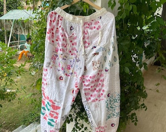 Puede incluir: Pantalones blancos de corte holgado con un diseño de patchwork que presenta varios patrones coloridos de flores, plumas de pavo real y formas geométricas. Los pantalones tienen una cintura elástica con cordón y cuelgan de una percha de madera.