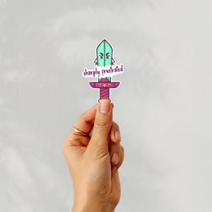 May include: A die-cut sticker featuring a cartoon sword with a mint green blade and a purple hilt. The sword has an angry facial expression and the text "sharply frustrated" written in purple script across the center of the blade.