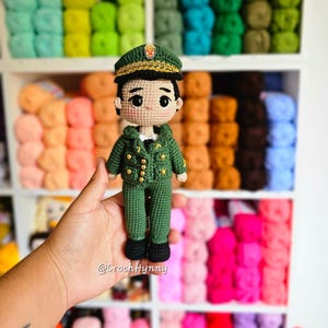 Puede incluir: Muñeca de ganchillo hecha a mano vestida con un uniforme verde de estilo militar con botones dorados y gorra a juego. La muñeca se sostiene frente a una estantería llena de madejas de hilo de colores. La cara de la muñeca tiene ojos negros y una expresión neutra.
