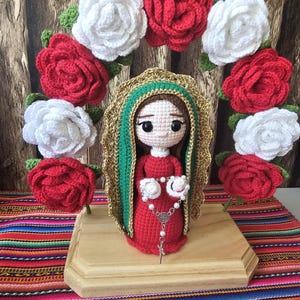 Peut inclure: Poupée au crochet faite à la main représentant la Vierge Marie, vêtue d'une robe rouge et d'un manteau vert et doré, ornée d'un chapelet de perles. La poupée est entourée d'une couronne de roses rouges et blanches au crochet, posée sur une base en bois.