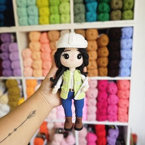 Op de afbeelding: Handgemaakte gehaakte pop van een bouwvakker, met een witte helm, groen vest, blauwe jeans en bruine laarzen. De pop houdt een wit papier vast. De achtergrond toont planken gevuld met kleurrijk garen.