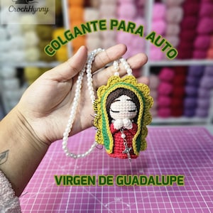 Może przedstawiać: Ręcznie robiony szydełkowy wisiorek do samochodu Virgen de Guadalupe. Wisiorek przedstawia Matkę Boską w kolorze czerwonym i zielonym, z perłowym naszyjnikiem i srebrnym krzyżem. Widoczny jest tekst "COLGANTE PARA AUTO" i "VIRGEN DE GUADALUPE".