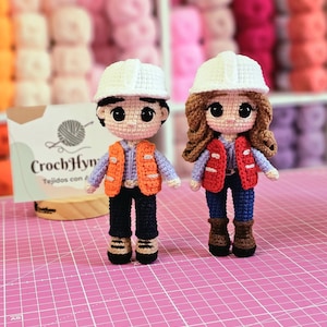 May include: Two handmade crochet dolls, one male and one female, dressed as construction workers. The male doll wears a white hard hat, orange vest, and blue pants. The female doll has brown hair, a red vest, and blue jeans. A sign reads "CrochHyn".
