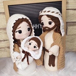 Puede incluir: Escena de la Natividad tejida a ganchillo con María, José y el niño Jesús. Las figuras están hechas de hilo blanco, marrón y beige. María sostiene al niño Jesús envuelto en una manta blanca. José está de pie junto a ellos, con una túnica beige.