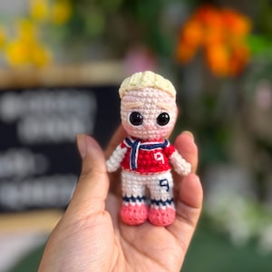 Puede incluir: Una pequeña muñeca de ganchillo con cabello rubio, que viste una camiseta deportiva roja y blanca con el número 9 en el pecho y pantalones cortos, una bufanda azul oscuro y zapatos rosas. La figura tiene grandes ojos negros y tejido detallado.