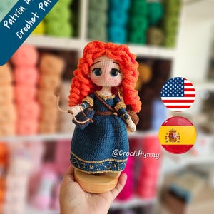 Può includere: Una bambola amigurumi all'uncinetto con vivaci capelli ricci arancioni, che indossa un abito blu scuro con finiture dorate, tenendo un piccolo arco e una freccia. La bambola ha grandi occhi blu e una base in legno. Testo Patrón Amigurumi Crochet Pattern.