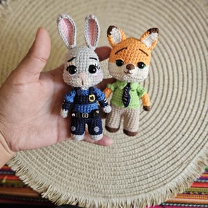 May include: Two crocheted dolls of characters from Zootopia. One is a gray bunny wearing a blue police uniform, and the other is an orange fox in a green shirt and tie. The dolls are handmade and have detailed features.