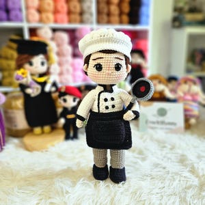 Puede incluir: Muñeco de chef tejido a ganchillo con gorro blanco, chaqueta y delantal negro, sosteniendo una pequeña sartén. El muñeco tiene una expresión neutra y está sobre una superficie blanca y esponjosa. Otros muñecos de ganchillo son visibles en el fondo.