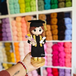 May include: A handmade crochet graduation doll in a black gown and mortarboard with a gold tassel. The doll holds a bouquet and a scroll. The doll is on a wooden base. The text "CrochHynny" is visible.