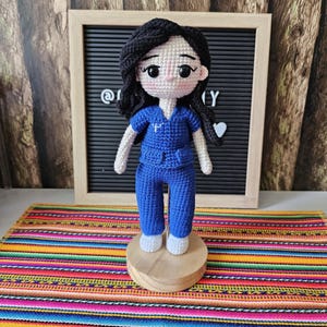 May include: A handmade crochet doll of a healthcare worker, wearing blue scrubs and white shoes. The doll has black hair and is standing on a wooden base. The background includes a framed letter board and a colorful striped textile.