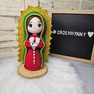Puede incluir: Muñeca de ganchillo hecha a mano de la Virgen María, con un vestido rojo, halo verde y dorado y un rosario plateado. La muñeca se exhibe sobre una base de rodaja de madera. El texto "@CROCHHYNNY" es visible sobre un fondo negro.